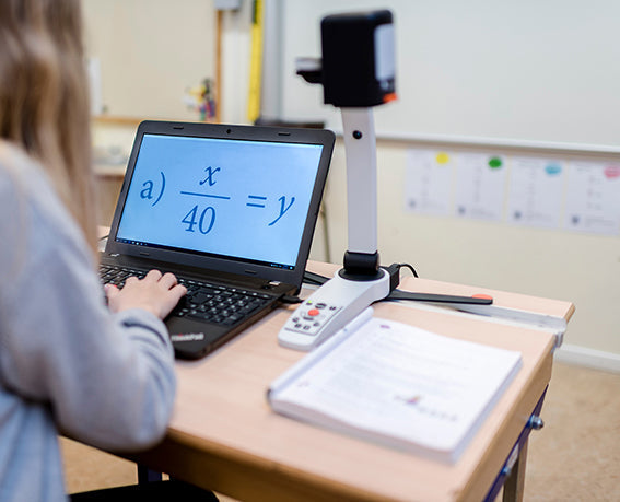 A student on a laptop magnified on a math problem from a book they have on their table, being viewed with a camera arm magnifier plugged into the laptop.