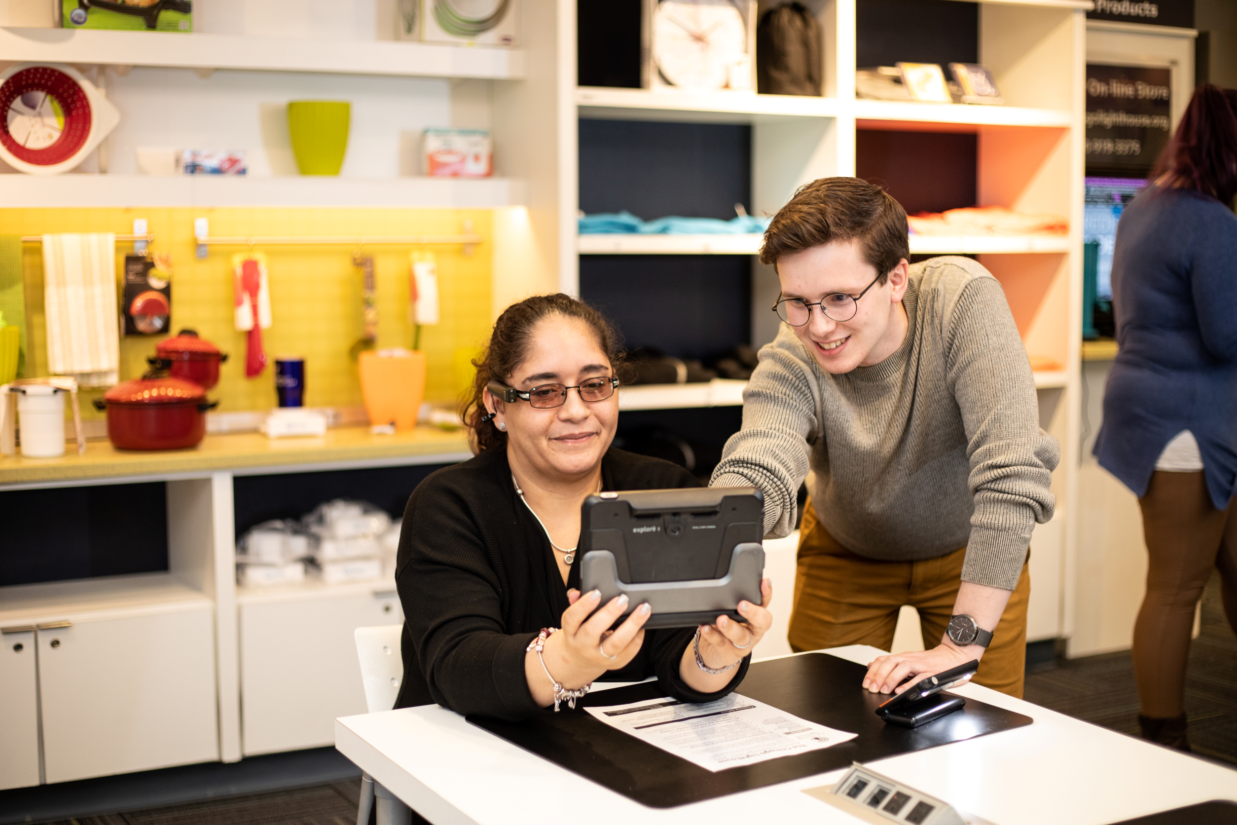 Two people together, one seated at the table utilizing a digital magnifier while wearing an OrCam while the other points at it.