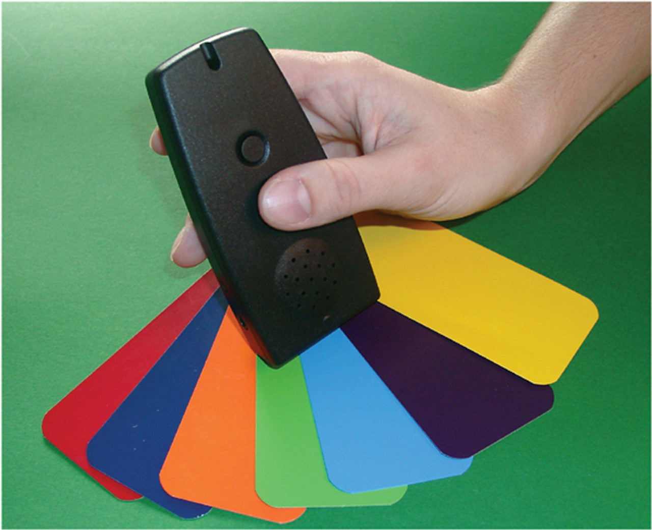 A black handheld device, the Colorino - Talking Color Identifier and Light Probe, is being held over several colorful rectangular cards. The cards, spread out like a fan, include bright hues such as red, orange, yellow, green, blue, and purple. The device has a small speaker grille and a circular button on the front, with a smooth and compact design. It is set against a green background.