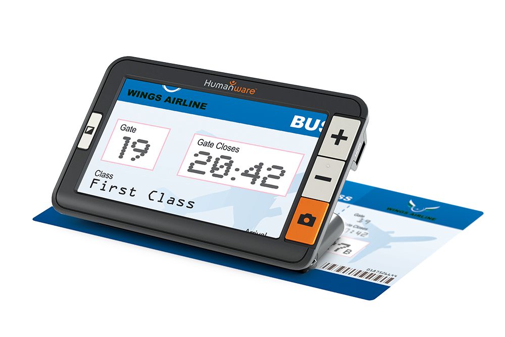 Humanware Explore 5 electronic magnifier which features a high definition LCD screen with physical buttons that include a bright orange camera big button and buttons to increase and decrease size. The device is angled and is sitting on a First Class Airline ticket to increase the size to make it easier to read. On the LCD screen is a large magnification of the ticket: the Gate number which is 19 and what time the Gate closes.