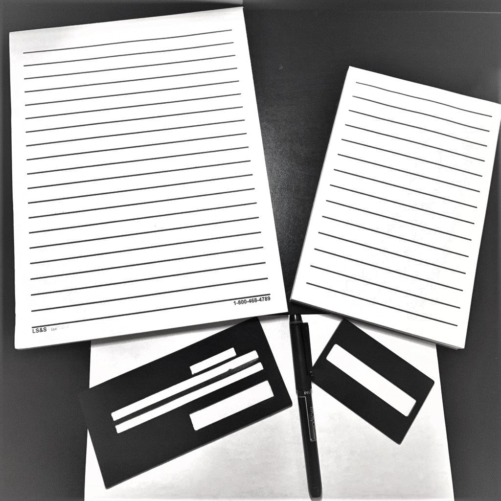 A bold line pad of paper and note pad with A checking writing guide, bravo pen, and aluminum signature guide below them